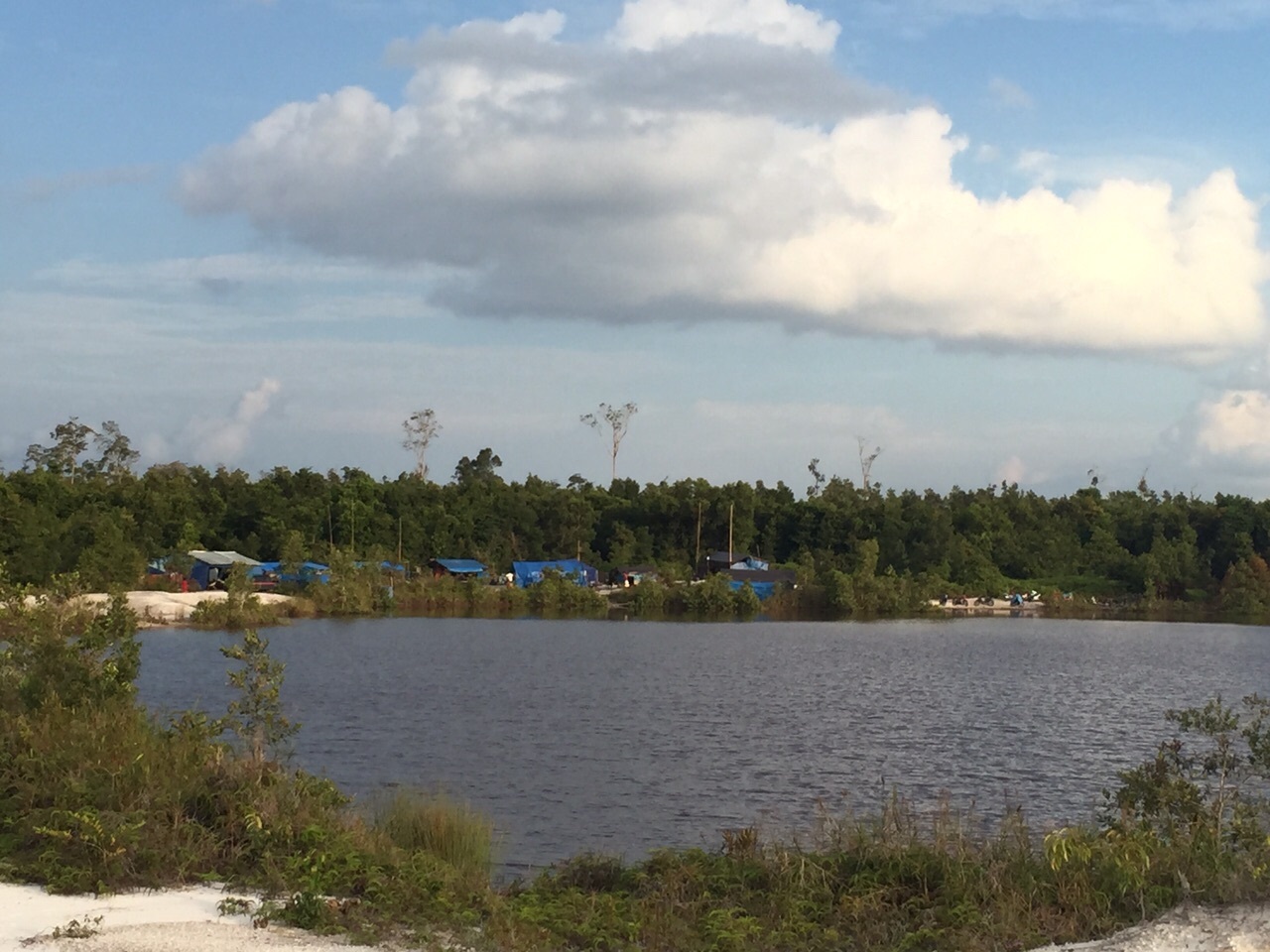 A view of a mine site with miners' tents in the background, next to a body of water but surrounded by barren land.